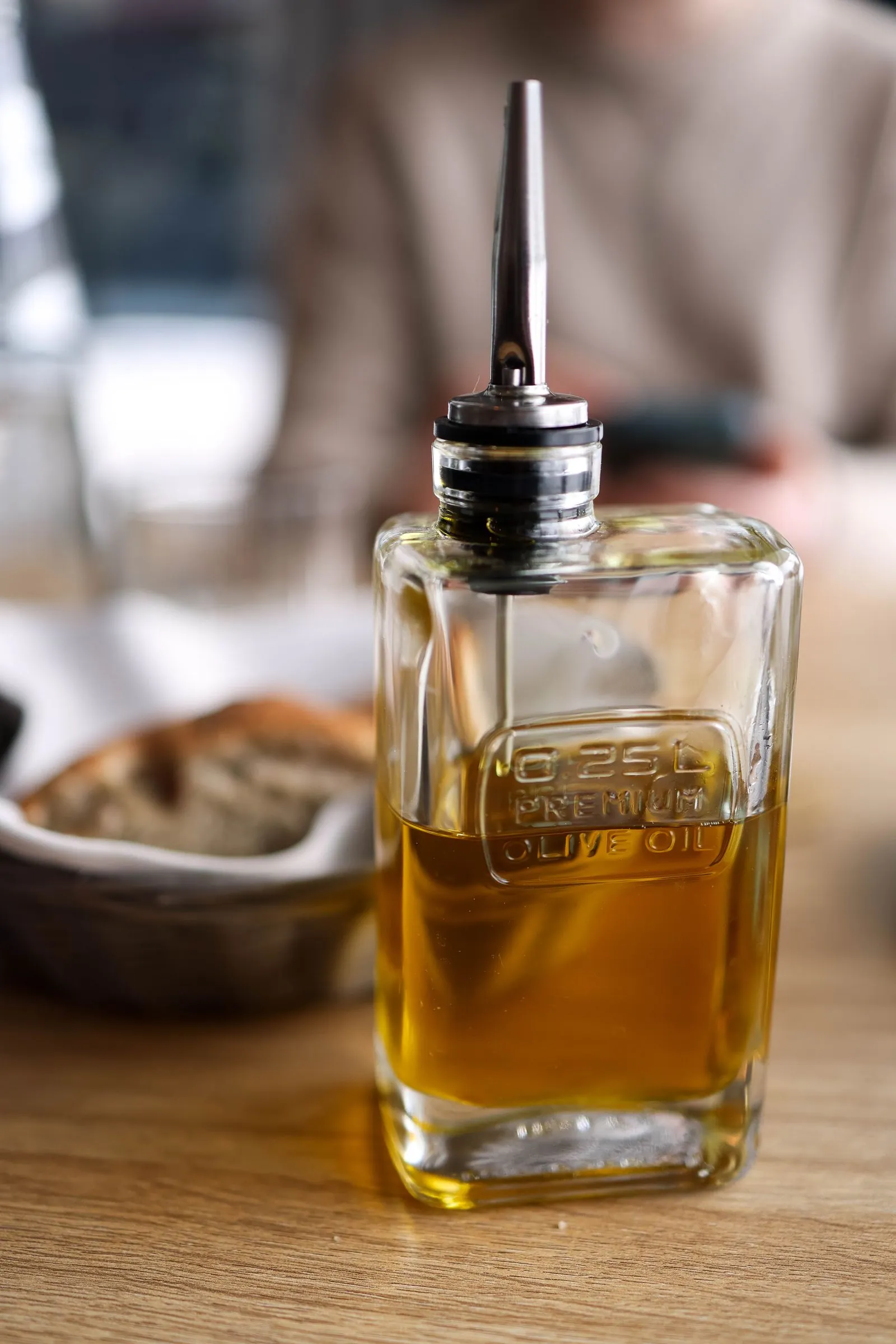 Illustrative photograph: An unlabelled clear glass oil bottle with a metal pouring spout on a wooden dining table.